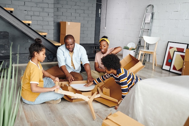 Jonge Afrikaanse familie zit op de vloer van een grote woonkamer in hun nieuwe flat of huis en haalt meubelonderdelen uit een kartonnen doos