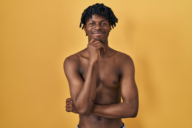 Jonge afrikaanse man met dreadlocks die shirtless staan en zelfverzekerd naar de camera kijken glimlachend met gekruiste armen en hand op kin. positief denken.