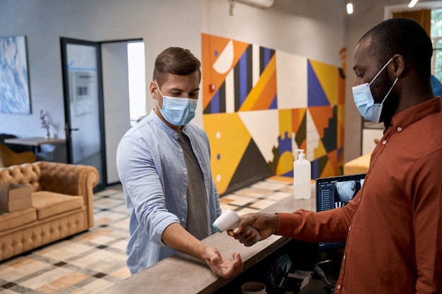 Jonge afrikaanse man met een medisch masker die bij de receptie in het moderne kantoor staat en meet