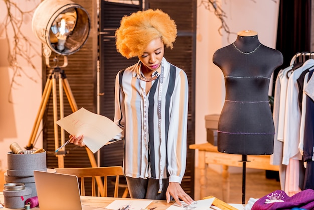Jonge afrikaanse modeontwerper die werkt met kledingtekeningen en laptop die in het interieur van de studio zit met verschillende kleermakers