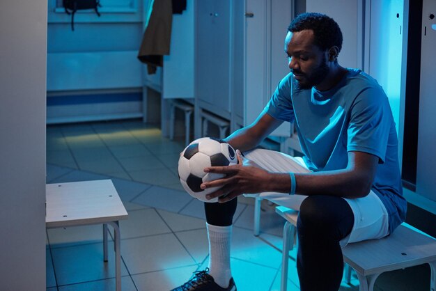 Jonge Afrikaanse sportman kijkt naar voetbal in zijn handen terwijl hij tijdens de pauze bij een kluisje in de kleedkamer zit