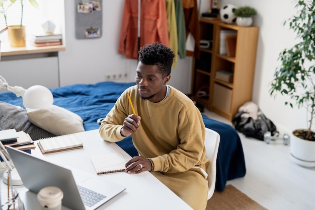 Jonge Afrikaanse student in homewear die voor laptop zit en naar leraar luistert tijdens online les