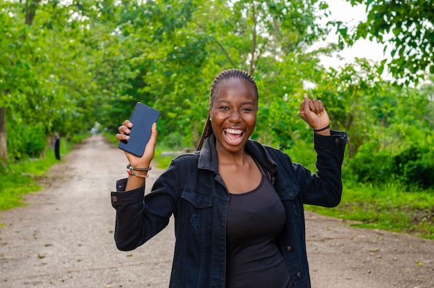 Foto jonge afrikaanse vrouw die buiten staat en opgetogen is tijdens het gebruik van hun smartphone