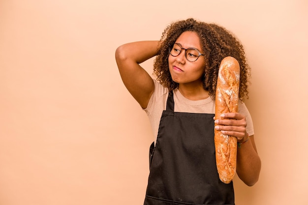 Jonge Afrikaanse vrouw die een brood vasthoudt, geïsoleerd op een beige achtergrond, achterhoofd aanraakt en een keuze maakt