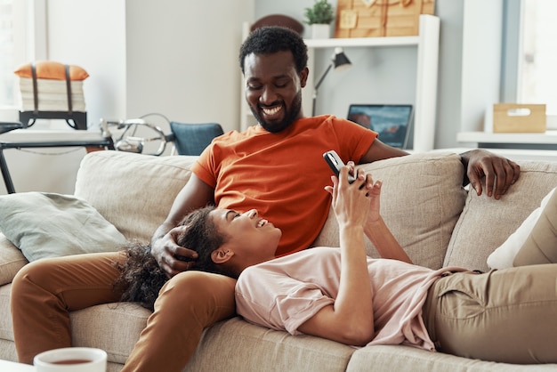 Jonge Afrikaanse vrouw die smartphone gebruikt en glimlacht terwijl ze thuis tijd doorbrengt met haar vriendje