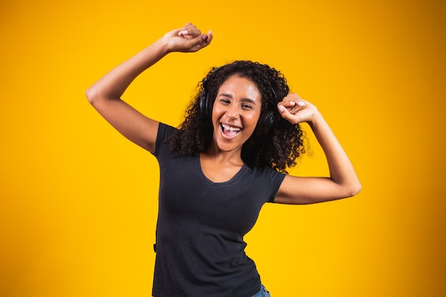 Jonge afrikaanse vrouw met koptelefoon die actief danst, geïsoleerd op gele muur, luisterend naar muziek die vrolijk lacht.