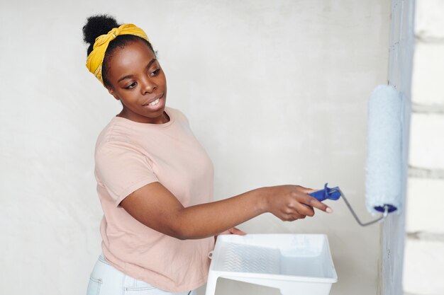 Jonge Afrikaanse vrouw met verfroller die voor de bakstenen muur van de woonkamer staat terwijl ze deze in witte kleur schildert tijdens de renovatie van het huis