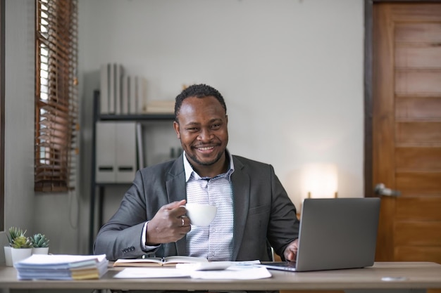 Jonge Afrikaanse zwarte mansmiling zakenman die koffie drinkt en laptop gebruikt in kantoor zittend aan een bureau