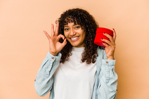 Jonge afro-amerikaanse afrovrouw die een vrolijke en zelfverzekerde Valentijnsdaggift houdt die ok gebaar toont.