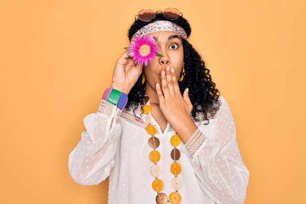 Foto jonge afro-amerikaanse gekrulde hippie-vrouw die een zonnebril draagt en roze bloem op de mond van de oogbedekking houdt met de hand geschokt van schaamte voor foute uitdrukking van angst bang in stilte geheim concept