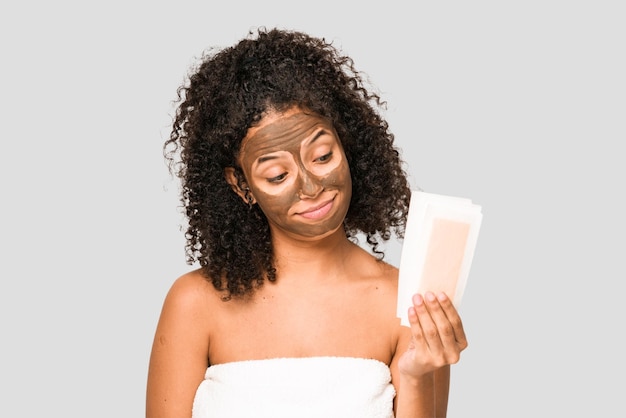 Jonge afro-amerikaanse gekrulde vrouw die voor zichzelf zorgt, ontharingsbanden vasthoudt en een geïsoleerd gezichtsmasker aanbrengt