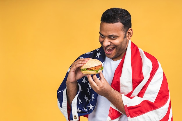Jonge Afro-Amerikaanse Indiase zwarte man eten hamburger geïsoleerd op gele achtergrond met Amerikaanse vlag. Dieet concept.