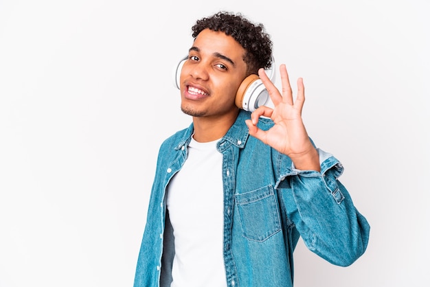 Jonge Afro-Amerikaanse krullende man geïsoleerd luisteren naar muziek met koptelefoon vrolijk en vol vertrouwen tonen ok gebaar.