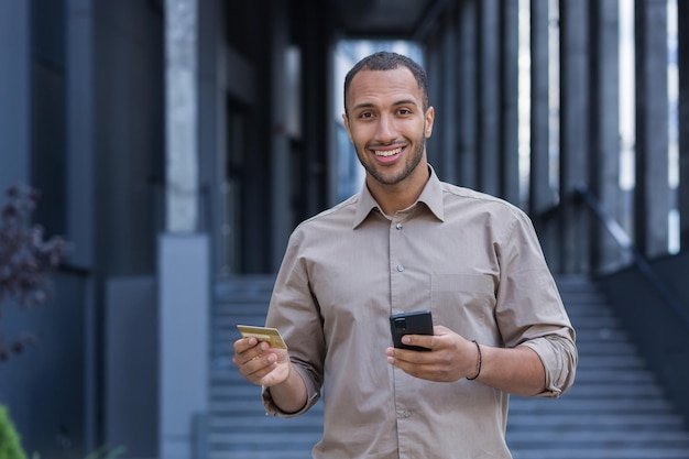 Jonge afro-amerikaanse man die buiten staat, hij houdt een telefoon en een creditcard in zijn handen maakt