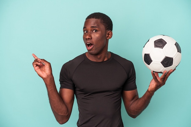 Jonge Afro-Amerikaanse man die voetbal speelt geïsoleerd op een blauwe achtergrond die naar de zijkant wijst