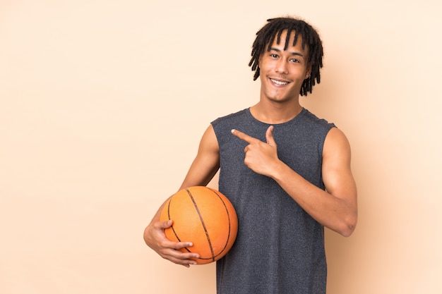 Jonge Afro-Amerikaanse man geïsoleerd op beige muur spelen basketbal en wijst naar de zijkant