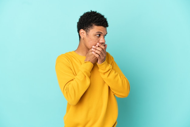 Jonge Afro-Amerikaanse man geïsoleerd op een blauwe achtergrond die de mond bedekt en naar de zijkant kijkt