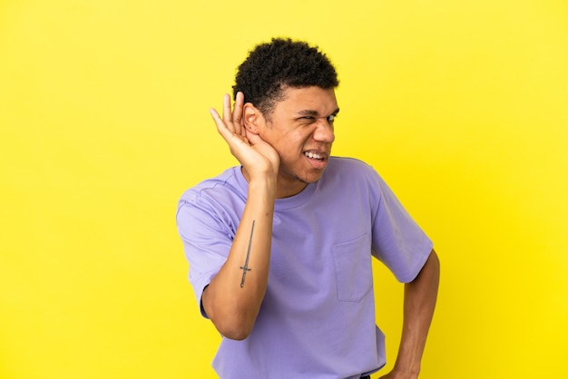 Jonge Afro-Amerikaanse man geïsoleerd op gele achtergrond luisteren naar iets door hand op het oor te leggen