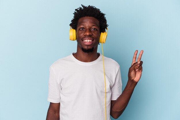 Jonge Afro-Amerikaanse man luisteren naar muziek geïsoleerd op blauwe achtergrond vrolijk en zorgeloos met een vredessymbool met vingers.
