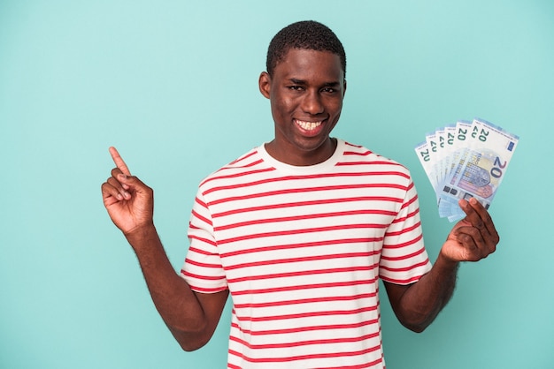 Jonge Afro-Amerikaanse man met bankbiljetten geïsoleerd op blauwe achtergrond glimlachend en opzij wijzend, iets latend op lege ruimte.