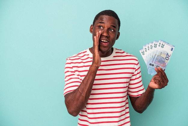 Jonge Afro-Amerikaanse man met bankbiljetten geïsoleerd op blauwe achtergrond zegt een geheim heet remnieuws en kijkt opzij