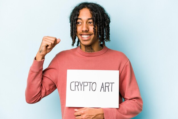 Jonge Afro-Amerikaanse man met crypto kunst plakkaat geïsoleerd op blauwe achtergrond die vuist opheft na een overwinning winnaar concept