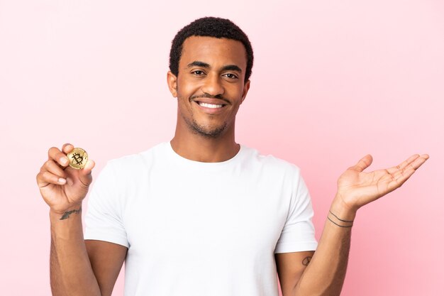 Jonge Afro-Amerikaanse man met een Bitcoin over een geïsoleerde roze achtergrond die zijn handen naar de zijkant uitstrekt om uit te nodigen om te komen