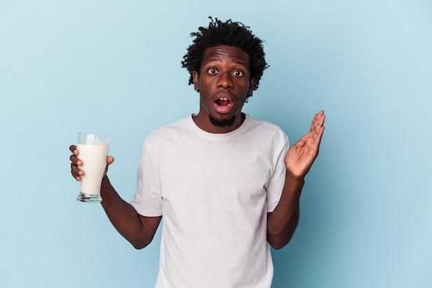 Jonge Afro-Amerikaanse man met een glas melk geïsoleerd op blauwe achtergrond verrast en geschokt.