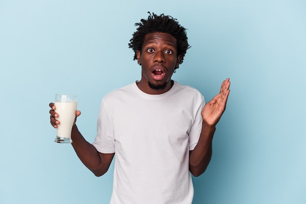 Jonge afro-amerikaanse man met een glas melk geïsoleerd op blauwe achtergrond verrast en geschokt.