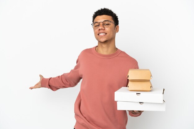 Jonge Afro-Amerikaanse man met een hamburger en pizza's geïsoleerd op een witte achtergrond die zijn handen naar de zijkant uitstrekt om uit te nodigen om te komen