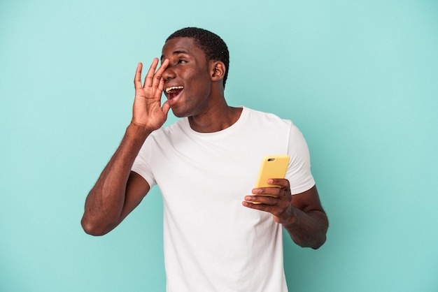 Jonge Afro-Amerikaanse man met een mobiele telefoon geïsoleerd op blauwe achtergrond schreeuwend en handpalm in de buurt van geopende mond.