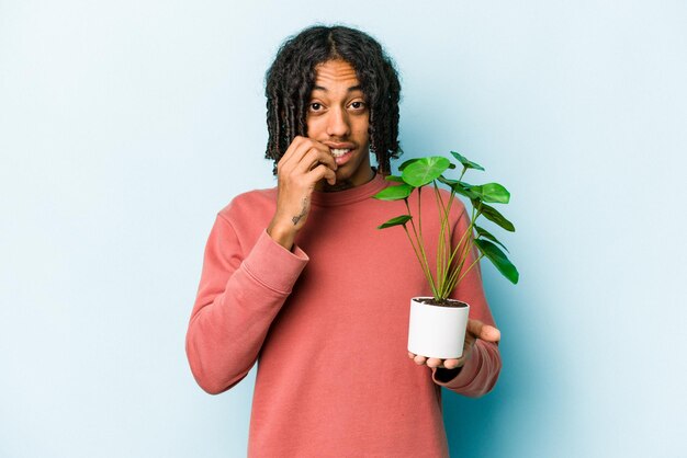 Jonge Afro-Amerikaanse man met een plant geïsoleerd op een blauwe achtergrond, nerveus en erg angstig vingernagels bijtend