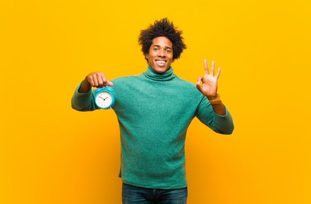 Jonge Afro-Amerikaanse man met een wekker tegen oranje ba