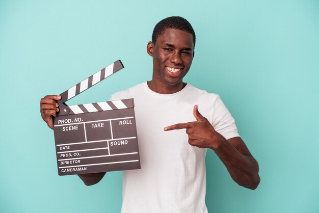 Jonge Afro-Amerikaanse man met Filmklapper geïsoleerd op blauwe achtergrond persoon wijzend met de hand naar een shirt kopie ruimte, trots en zelfverzekerd