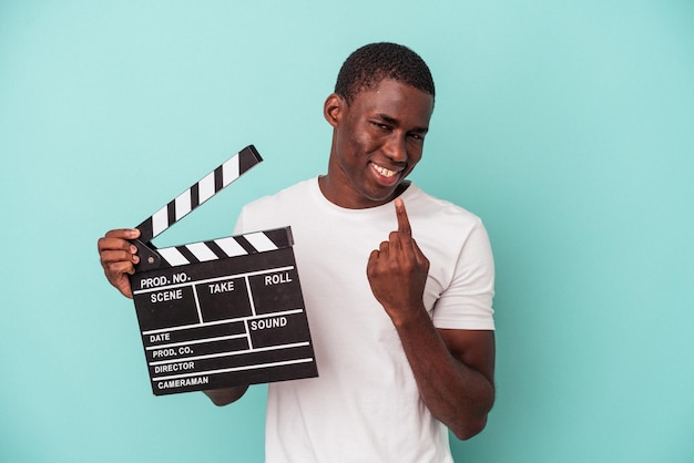 Jonge afro-amerikaanse man met filmklapper geïsoleerd op blauwe achtergrond wijzend met de vinger naar je alsof uitnodigend dichterbij komt.