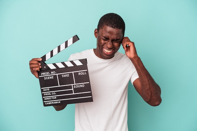 Jonge Afro-Amerikaanse man met Filmklapper geïsoleerd op een blauwe achtergrond die oren bedekt met handen.