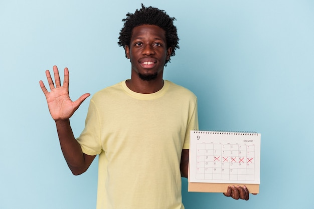 Jonge Afro-Amerikaanse man met kalender geïsoleerd op blauwe achtergrond glimlachend vrolijk nummer vijf met vingers.