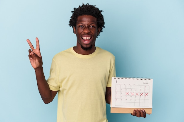 Jonge Afro-Amerikaanse man met kalender geïsoleerd op blauwe achtergrond vrolijk en zorgeloos met een vredessymbool met vingers.