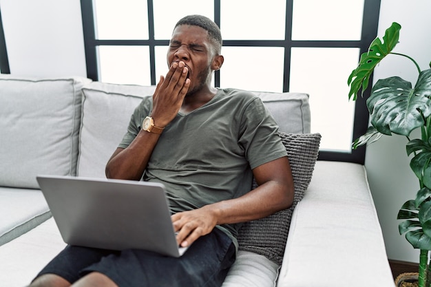 Jonge afro-amerikaanse man met laptop thuis zittend op de bank verveeld geeuwen moe bedekkende mond met hand rusteloos en slaperigheid