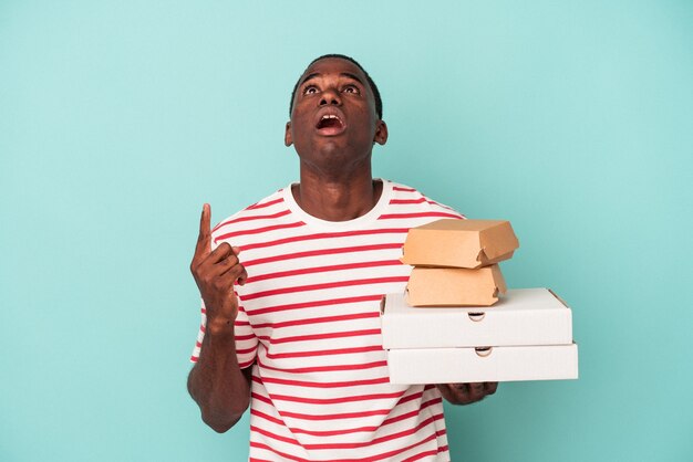 Jonge Afro-Amerikaanse man met pizza's en hamburgers geïsoleerd op blauwe achtergrond wijzend ondersteboven met geopende mond.