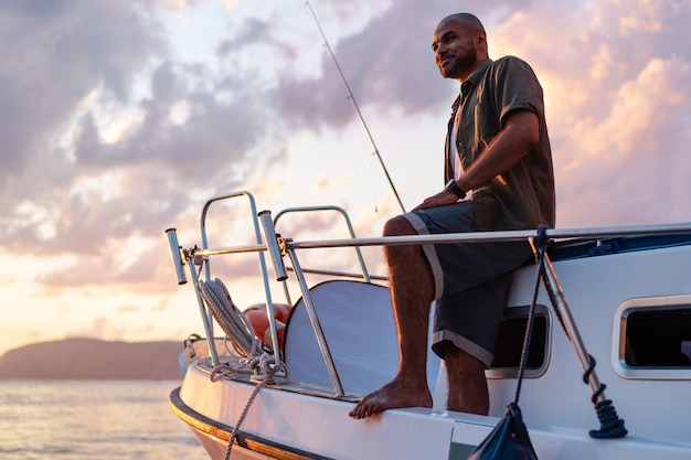 Jonge Afro-Amerikaanse man permanent met hengel op een zeilboot vissen in open zee op zonsondergang