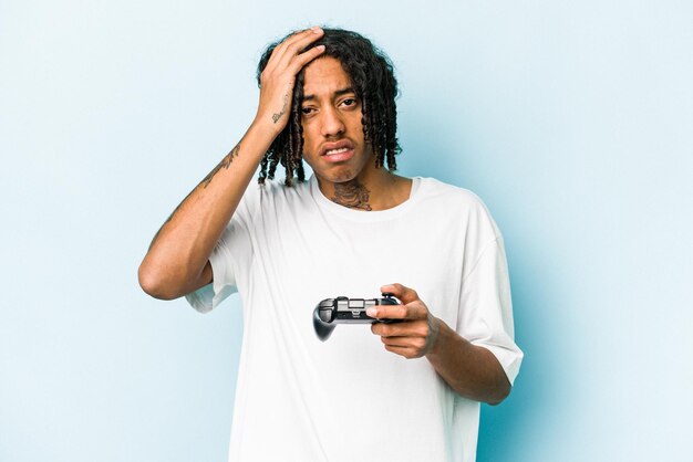 Jonge Afro-Amerikaanse man speelt met een videogamecontroller geïsoleerd op een blauwe achtergrond en is geschokt dat ze zich een belangrijke ontmoeting heeft herinnerd