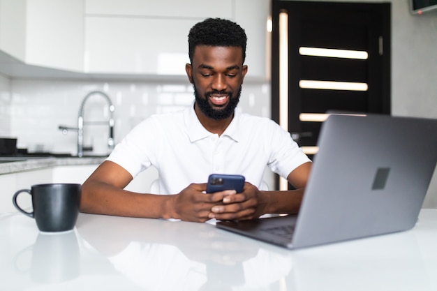 Jonge Afro-Amerikaanse man telefoon gebruiken zittend met laptop in de keuken