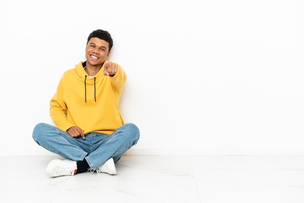 Jonge Afro-Amerikaanse man zittend op de vloer geïsoleerd op een witte achtergrond wijst vinger naar je met een zelfverzekerde uitdrukking