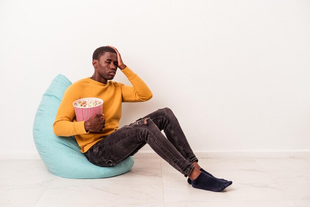 Jonge Afro-Amerikaanse man zittend op een trekje popcorn eten geïsoleerd op een witte achtergrond, geschokt, ze heeft een belangrijke ontmoeting onthouden