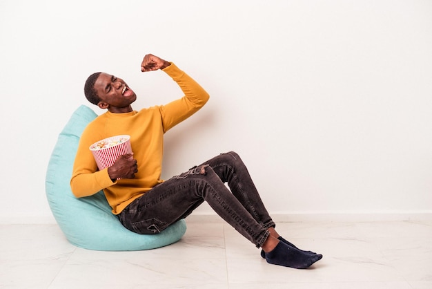 Jonge Afro-Amerikaanse man zittend op een trekje popcorn eten geïsoleerd op een witte achtergrond vuist opheffen na een overwinning, winnaar concept.