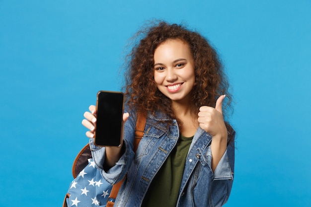 Jonge Afro-Amerikaanse meisje tiener student in denim kleding, rugzak houden telefoon geïsoleerd op blauwe muur