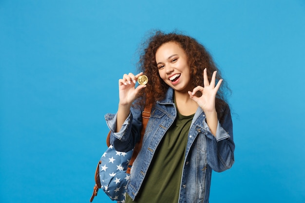 Jonge Afro-Amerikaanse meisje tiener student in denim kleding, rugzak houdt bitcoin geïsoleerd op blauwe muur