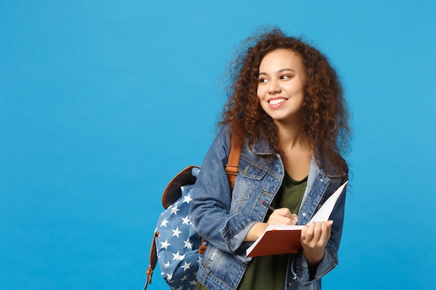 Jonge Afro-Amerikaanse meisje tiener student in denim kleding, rugzak houdt boeken geïsoleerd op blauwe muur