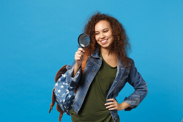 Jonge Afro-Amerikaanse meisje tiener student in denim kleding, rugzak houdt klok geïsoleerd op blauwe muur